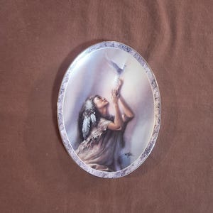 May include: Oval decorative plate featuring a Native American woman with a feathered headdress, reaching towards a white dove. The plate has a silver-toned border with a detailed design. The artwork is set against a soft, diffused background.