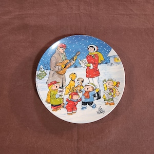 May include: A decorative plate featuring a winter scene with people singing carols. A man plays a guitar, and a woman holds a songbook. Children in winter attire and dogs are also present. The plate has a blue and white background with falling snow.