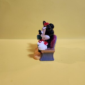 Pu&ograve; includere: Una statuina di Minnie Mouse, con testa nera e fiocco rosso, guanti bianchi e un vestito a pois rossi e bianchi. La statuina &egrave; seduta su una sedia viola e marrone, che tiene in mano una scatola regalo verde.