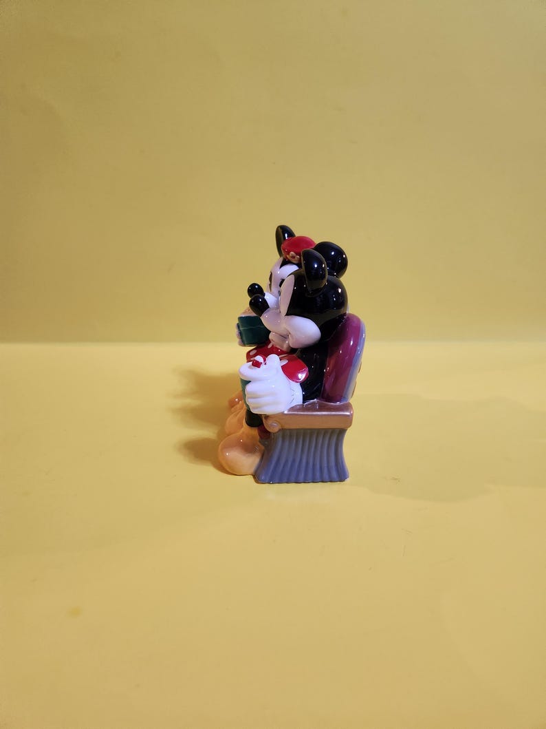 Pu&ograve; includere: Una statuina di Minnie Mouse, con testa nera e fiocco rosso, guanti bianchi e un vestito a pois rossi e bianchi. La statuina &egrave; seduta su una sedia viola e marrone, che tiene in mano una scatola regalo verde.