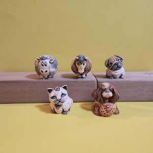 May include: Five small animal figurines: a cow, a horse, a pug, a cat, and a dog. The cow is white and gray, the horse is brown and tan, the pug is tan and white, the cat is white with blue eyes, and the dog is brown and tan.