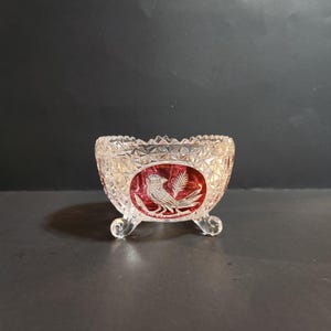 May include: A clear, footed glass bowl with a red oval design featuring a bird and wheat. The bowl has a diamond-cut pattern around the rim and on the sides. The bowl is supported by three small, rounded feet.