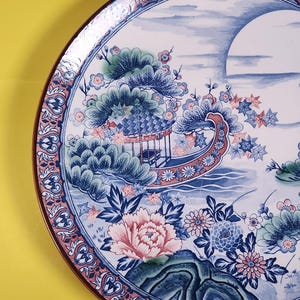 May include: Decorative ceramic plate with a Japanese-inspired design. The plate features a detailed illustration of a boat, flowers, and a landscape in shades of blue, pink, and green, with a brown rim. The plate's diameter is approximately 30 cm.