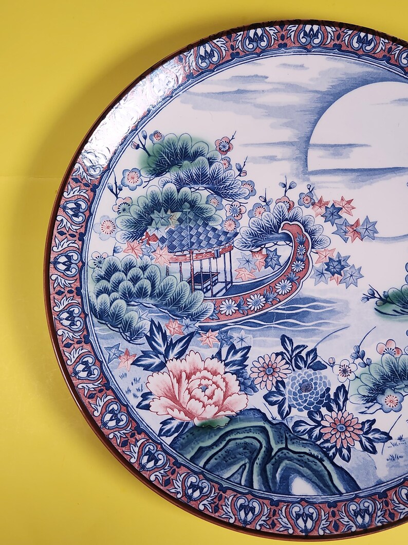 May include: Decorative ceramic plate with a Japanese-inspired design. The plate features a detailed illustration of a boat, flowers, and a landscape in shades of blue, pink, and green, with a brown rim. The plate's diameter is approximately 30 cm.