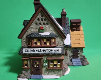 May include: A small, detailed ceramic house with a brown roof and white snow accents. The house has a sign that reads "Giggelswick-Mutton & Ham".