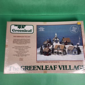 May include: A Greenleaf Village model kit box featuring a photograph of six miniature wooden houses and a church arranged in a snowy landscape. The packaging includes the brand name, product title, assembly details, and a toll-free phone number.