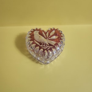 May include: A heart-shaped glass trinket box with a clear glass lid and a red and white etched design of a bird with a leafy branch.