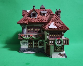 Puede incluir: Un edificio blanco en miniatura con un techo rojo y un letrero que dice "The Lechfield Solicitor". El edificio tiene un balcón con una corona verde y un techo cubierto de nieve.