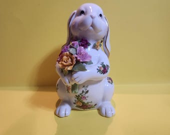 May include: A white ceramic bunny figurine holding a bouquet of flowers. The bunny has floral accents and gold trim on its ears. The flowers are in shades of pink, purple, and orange. The figurine is on a yellow background.