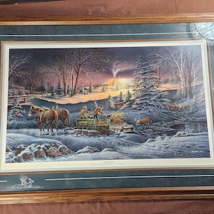May include: Framed winter landscape print featuring a horse-drawn sleigh carrying people through a snowy scene. The artwork depicts a cabin, trees, and deer, with a warm sunset glow. The frame is wood with a green matting.