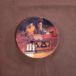 May include: A decorative plate featuring a vintage illustration with a boxer, a man in a suit, and a tea box. The plate also depicts an I.G.A. Stores building and the text "Emperor Hirohito". The plate has a gold rim.