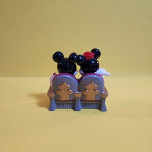 Pu&ograve; includere: Una statuetta di Topolino e Minnie seduti su una poltrona teatrale blu e oro. Topolino indossa un papillon nero e Minnie indossa un fiocco rosso. Lo sfondo &egrave; giallo pieno.