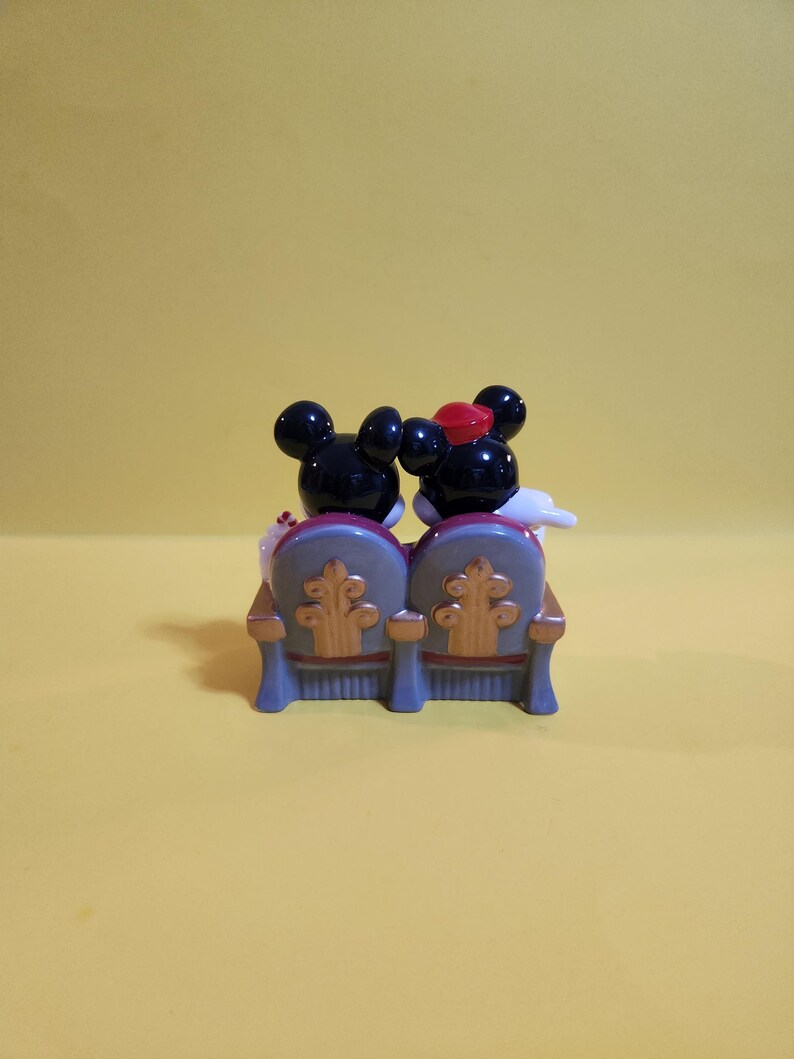 Pu&ograve; includere: Una statuetta di Topolino e Minnie seduti su una poltrona teatrale blu e oro. Topolino indossa un papillon nero e Minnie indossa un fiocco rosso. Lo sfondo &egrave; giallo pieno.