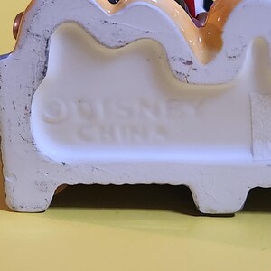 Pu&ograve; includere: Statuetta in ceramica con base bianca e dettagli colorati. La base presenta la scritta in rilievo "DISNEY CHINA". La statuetta raffigura un personaggio dei cartoni animati con un abito rosso, bianco e blu.