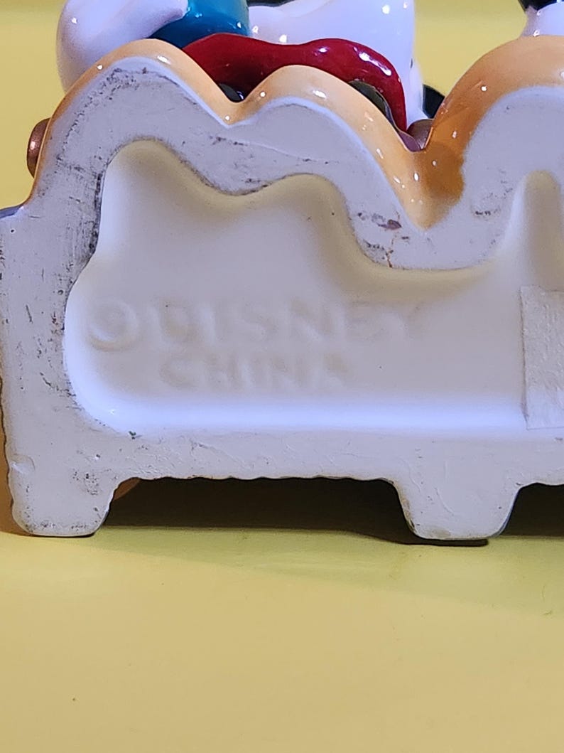 Pu&ograve; includere: Statuetta in ceramica con base bianca e dettagli colorati. La base presenta la scritta in rilievo "DISNEY CHINA". La statuetta raffigura un personaggio dei cartoni animati con un abito rosso, bianco e blu.