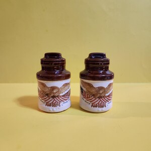 May include: Two brown and white ceramic bottles with a dark brown top. Each bottle features a design of an eagle with outstretched wings over a red and white striped background. The text "The Spirit of Liberty" is printed on the bottom.