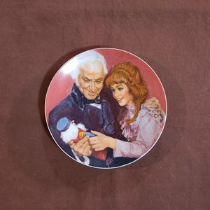 May include: A decorative plate featuring a painted scene of an older man and a young woman looking at an object. The plate has a red border and a glossy finish. The man is wearing a dark coat, and the woman is wearing a pink dress.