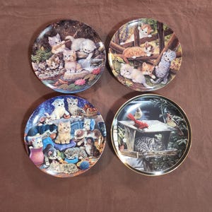 May include: Four decorative plates featuring detailed illustrations of kittens in various settings. One plate shows kittens near a stream, another with kittens on a fence, a third with kittens in a room, and the last with a sleeping cat near a bird feeder.