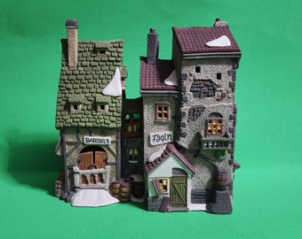 May include: A ceramic village scene depicting three connected buildings with textured roofs in green, red, and grey. The building on the left has a sign that reads "BARRELS". The middle building has a sign that reads "Faqin". Snow accents the roofs, creating a wintery scene.