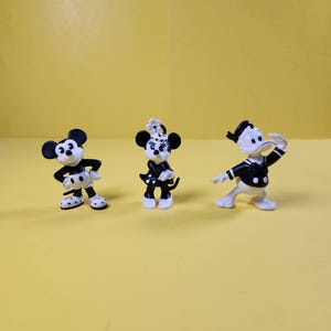 May include: Three small, black and white cartoon character figurines standing in a row. The left figure has large round ears, the center figure wears a polka-dot dress and bow, and the right figure is a duck wearing a sailor-style shirt and cap.