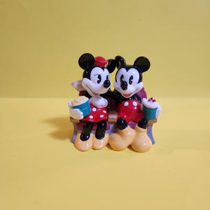 Pu&ograve; includere: Statuina in ceramica di Topolino e Minnie seduti su una panchina. Minnie indossa un vestito rosso a pois bianchi e un cappello rosso. Topolino indossa pantaloncini rossi. Entrambi tengono popcorn e bevande.