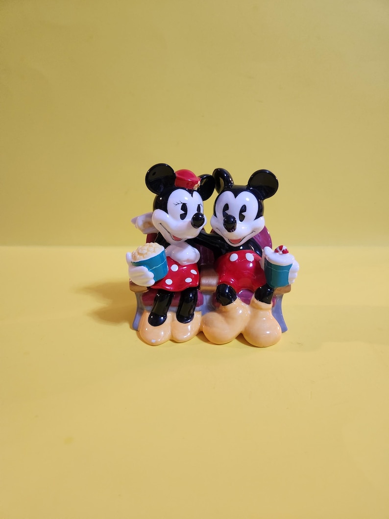 Pu&ograve; includere: Statuina in ceramica di Topolino e Minnie seduti su una panchina. Minnie indossa un vestito rosso a pois bianchi e un cappello rosso. Topolino indossa pantaloncini rossi. Entrambi tengono popcorn e bevande.
