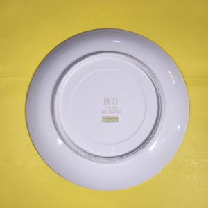 May include: Underside view of a white ceramic plate with a broad rim. The centre of the plate has a smaller, recessed area. The words "HAND DECORATED" are printed on a small label. The plate is set against a yellow background.