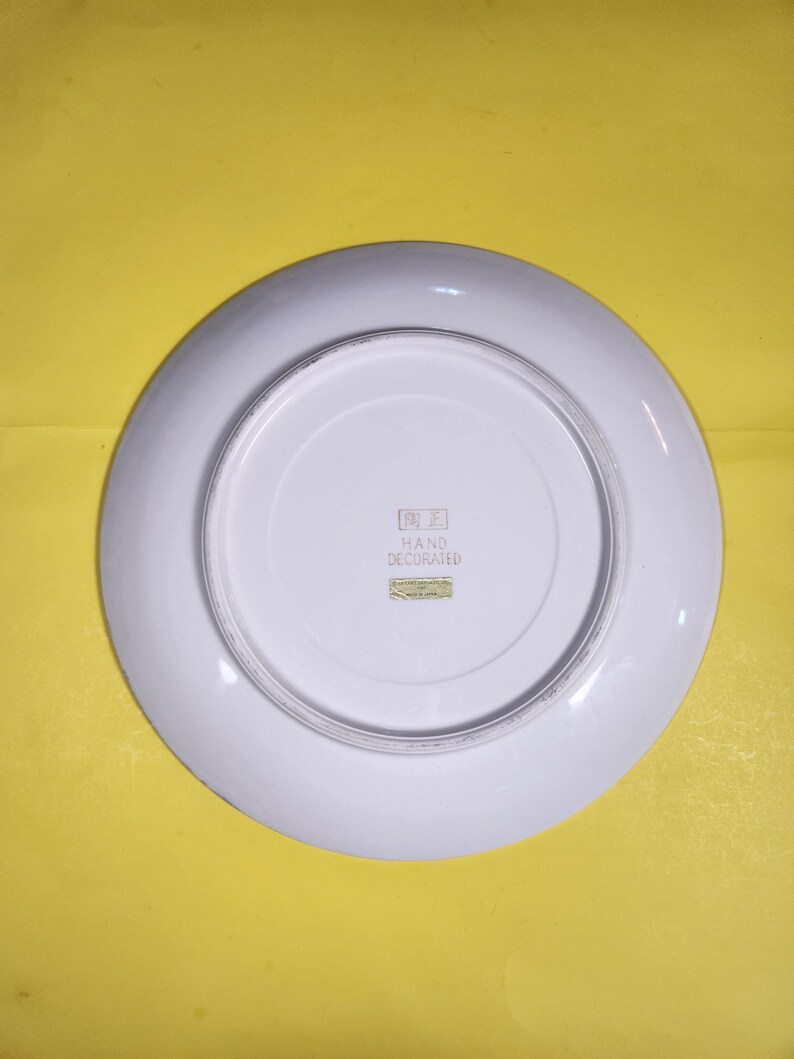 May include: Underside view of a white ceramic plate with a broad rim. The centre of the plate has a smaller, recessed area. The words "HAND DECORATED" are printed on a small label. The plate is set against a yellow background.