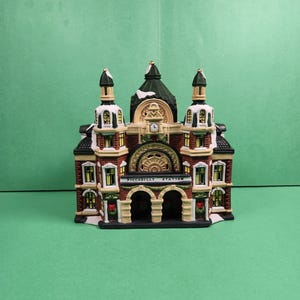 May include: A ceramic Piccadilly Station miniature with a detailed facade. The building features red brick, white trim, and a green roof. The text "PICCADILLY STATION" is displayed above the arched entrance. The miniature has three towers and a clock.