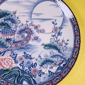 May include: A decorative ceramic plate with an Asian-inspired design. The plate features a blue and white landscape with a large moon, flowers, and a pagoda. The rim has a red and blue patterned border.