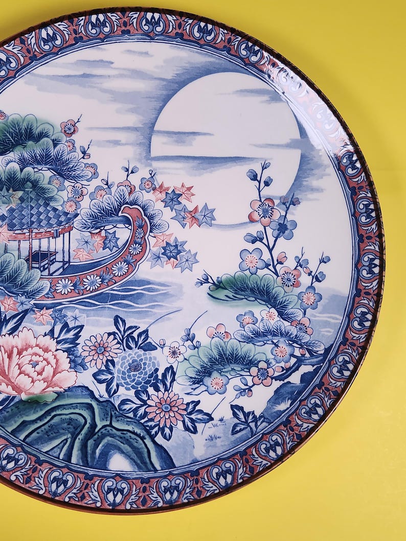 May include: A decorative ceramic plate with an Asian-inspired design. The plate features a blue and white landscape with a large moon, flowers, and a pagoda. The rim has a red and blue patterned border.