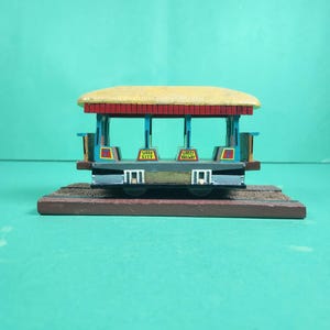 May include: A miniature model of a Sioux City Street Railway trolley car featuring a yellow roof, red trim, and blue structural supports. The open-air vehicle displays the number 7 on its sides and sits on a brown rectangular track base.