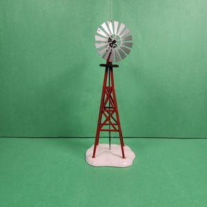 May include: A miniature red and silver windmill with a white base. The windmill features a silver fan and a red support structure. The base is white and shaped like a snowdrift. The windmill is set against a green background.