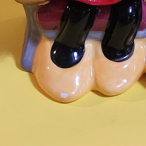 Pu&ograve; includere: Una statuetta in ceramica raffigurante le gambe e l'abito di un personaggio. L'abito &egrave; rosso a pois bianchi. Le scarpe del personaggio sono nere e lucide. La statuetta ha una base beige su sfondo giallo.