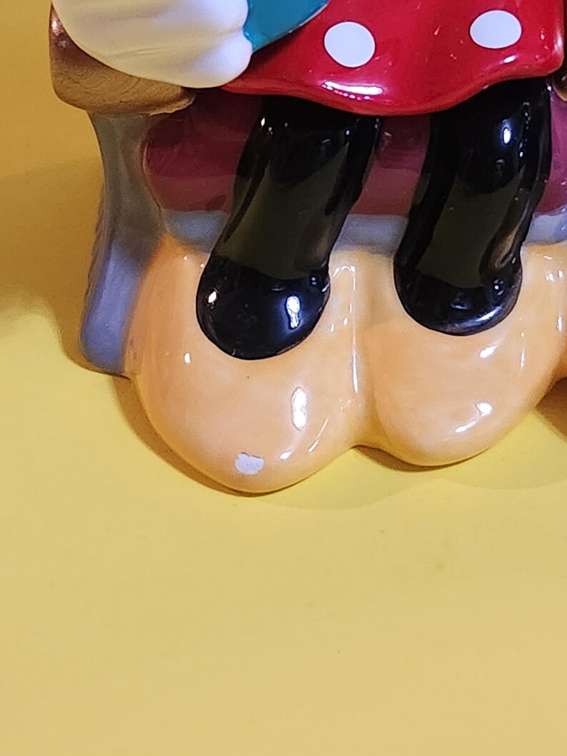 Pu&ograve; includere: Una statuetta in ceramica raffigurante le gambe e l'abito di un personaggio. L'abito &egrave; rosso a pois bianchi. Le scarpe del personaggio sono nere e lucide. La statuetta ha una base beige su sfondo giallo.