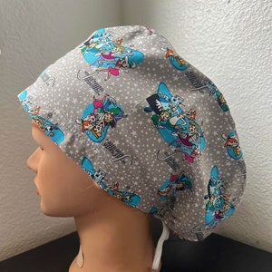 May include: Gray surgical scrub cap featuring a cartoon design with characters in blue circles and white stars. The word "Justice" is written in cursive. The cap has a white drawstring for adjustment.