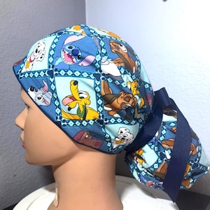 May include: Blue scrub cap featuring cartoon characters like Stitch, Pluto, and Simba. The cap has a dark blue ribbon and is designed with a repeating pattern of squares. The background is a light blue color.