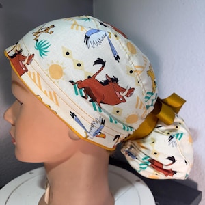 May include: A white scrub cap featuring a repeating pattern of cartoon characters, including a warthog, giraffe, and bird, with yellow suns and geometric shapes. The cap has a yellow trim and a matching bow.