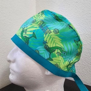May include: A turquoise blue and green patterned surgical scrub hat with a turquoise blue ribbon. The hat has a tropical print with palm leaves, green and yellow limes, and martini glasses. The text "It's 5 o'clock somewhere" is printed on the hat.
