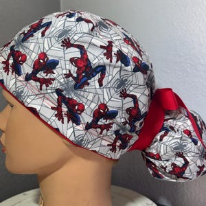 May include: A white and grey scrub cap featuring a Spider-Man design with red and blue figures and spiderwebs. The cap has a red trim and a red bow. The cap is designed to be worn on the head.