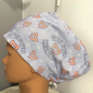 Puede incluir: Un gorro quirúrgico azul claro con un patrón repetido de intestinos naranjas y formas florales blancas. El texto en el gorro dice "GENERAL SURGERY" y "WE'RE A PAIN IN THE BUTT!"