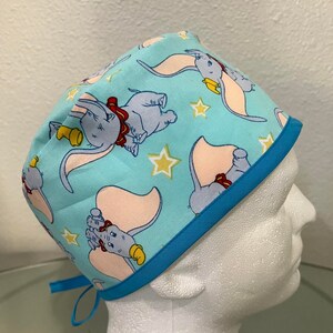May include: A blue and turquoise scrub hat with a pattern of Dumbo the elephant and stars. The hat has a turquoise trim and ties.