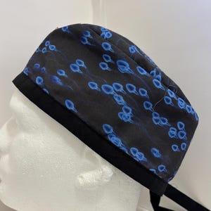 May include: A black surgical scrub cap with a repeating blue sperm cell pattern. The cap has a black band around the bottom and black ties. The cap is designed to be worn in a medical setting.