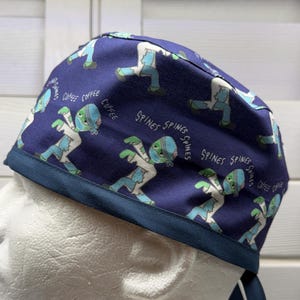 May include: A dark blue scrub cap featuring a repeating pattern of cartoon zombies wearing medical coats and face masks. The fabric includes the printed words COFFEE and SPINES scattered among the zombie figures, finished with a solid dark blue band.