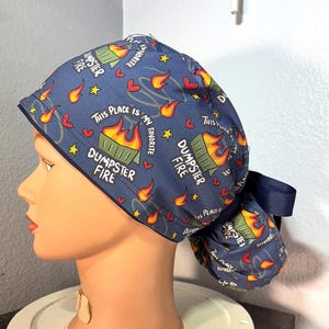 May include: A navy blue scrub cap with a repeating pattern of burning dumpsters, flames, hearts, and stars. The text "THIS PLACE IS MY FAVORITE" and "DUMPSTER FIRE" are also printed on the cap. A dark blue ribbon is attached.