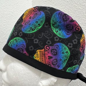 Puede incluir: Un gorro de quirófano negro con un patrón de calavera de azúcar arcoíris. El gorro tiene una banda elástica negra y lazos.