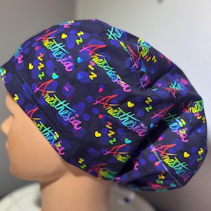 Könnte beinhalten: Eine marineblaue OP-Haube mit einem bunten Muster. Die Haube zeigt das Wort "Anesthesia" in Regenbogen-Schrift, zusammen mit Herzen, Z's und Punkten. Die Haube ist für medizinisches Fachpersonal bestimmt.