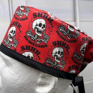 May include: A red scrub cap featuring a repeating pattern of white skeletons with black outlines, each holding a finger to their mouth. The text "SHHH" and "NO ONE CARES" is printed in black around the skeletons. The cap has a solid black fabric band.