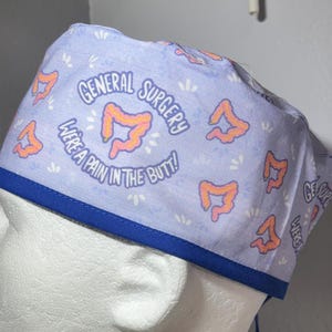 Pode incluir: Uma touca cirúrgica roxa clara com acabamento azul. A touca apresenta o texto "GENERAL SURGERY WE'RE A PAIN IN THE BUTT!" e ilustrações laranja de intestinos. A touca foi concebida para ser usada em ambiente médico.