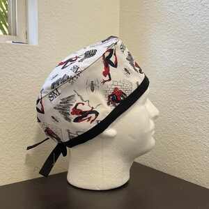 Peut inclure: Un bonnet de travail blanc avec une bordure noire et un motif répété de personnages Spiderman en rouge et noir. Le bonnet a des liens noirs.